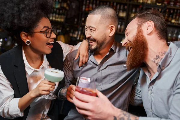 Возбужденные мультиэтнические коллеги с напитками, обнимающие и смеющиеся в баре, досуг после работы — стоковое фото