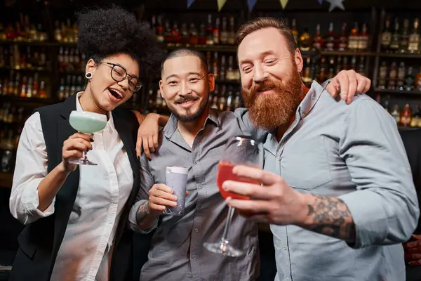Barbuto asiatico uomo guardando fotocamera vicino allegro multietnico amici con cocktail in bar — Foto stock