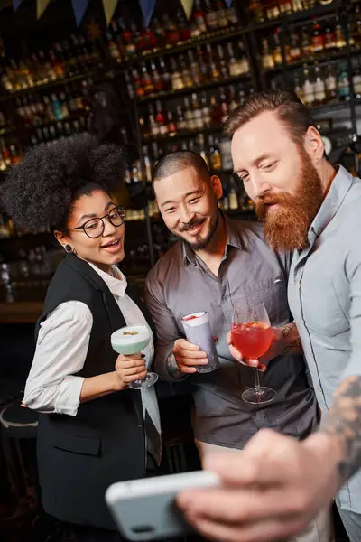 Бородатый мужчина делает селфи на смартфоне с счастливыми мультиэтническими коллегами, держащими коктейли в баре — стоковое фото
