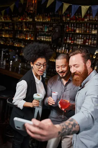 Tattooed bearded man taking selfie on smartphone with smiling multiethnic colleagues in cocktail bar — Stock Photo