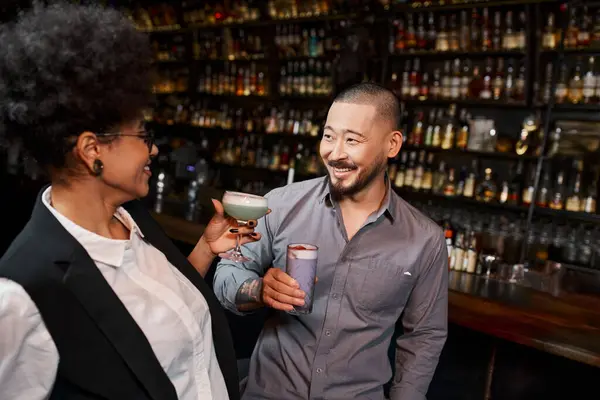 Joyful asian man and african american woman holding cocktails glasses and talking in bar after work — Stock Photo