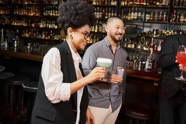 Азіатський чоловік і афроамериканська жінка тримає коктейлі і посміхається під час після робочої вечірки в барі — стокове фото