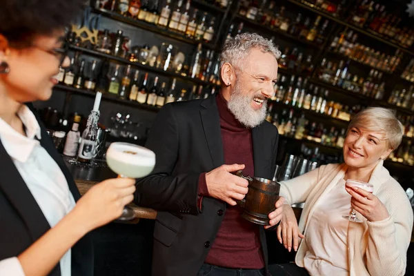 Multiethnic women holding glasses and looking at cheerful bearded colleague in cocktail bar — Stock Photo