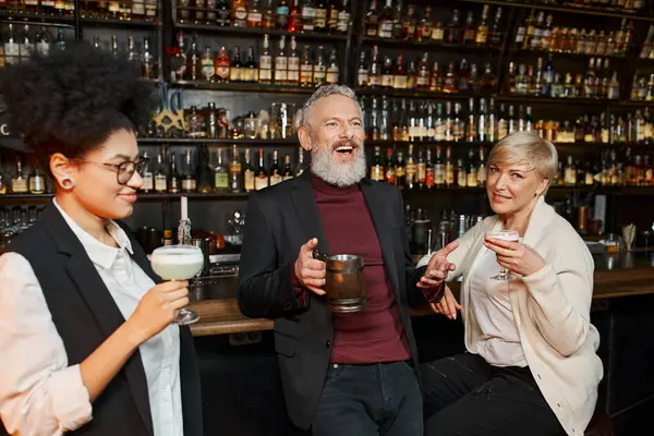 Веселый бородатый мужчина смеется рядом с многонациональными женщинами в коктейльных бокалах в баре, после рабочей вечеринки — стоковое фото
