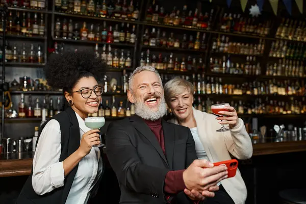 Homme barbu joyeux prenant des photos avec des femmes multiethniques dans le bar, équipe diversifiée se reposant après le travail — Photo de stock