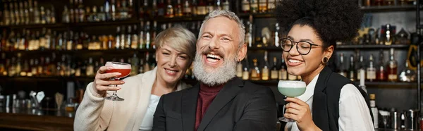 Happy multiethnic women holding cocktail glasses near bearded colleague laughing in bar, banner — Stock Photo