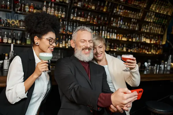 Joyful multiethnic women with cocktails near bearded colleague taking selfie on smartphone in bar — Stock Photo