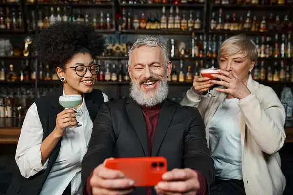 Щасливий бородатий чоловік бере селфі на смартфон з мультикультурними жінками з коктейль-окулярами в барі — стокове фото