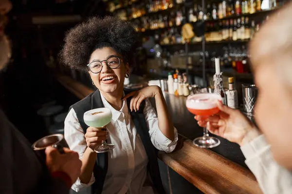 Femme afro-américaine gaie tenant verre à cocktail tout en passant du temps avec des collègues dans le bar — Photo de stock
