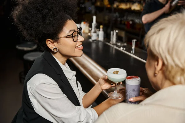 Happy african american woman with blurred female colleague holding glasses with cocktails in bar — Stock Photo