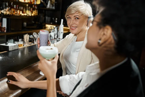 Happy middle aged woman with female african american colleague drinking cocktails in bar after work — Stock Photo