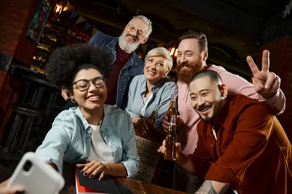 Femme afro-américaine gaie prenant des photos avec des collègues multiethniques tenant des bouteilles de bière dans un pub — Photo de stock