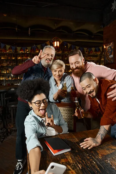 Mulher afro-americana mostrando sinal de rocha enquanto toma selfie com colegas de trabalho multiétnicos no pub — Fotografia de Stock