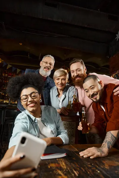 Joyful african american woman showing rock sign and taking selfie with multiethnic workmates in pub — Stock Photo