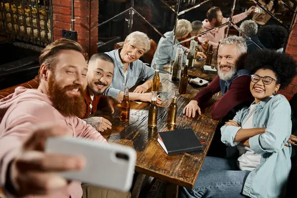 Happy bearded man taking selfie with multiethnic colleagues near beer bottles on wooden table in pub — Stock Photo