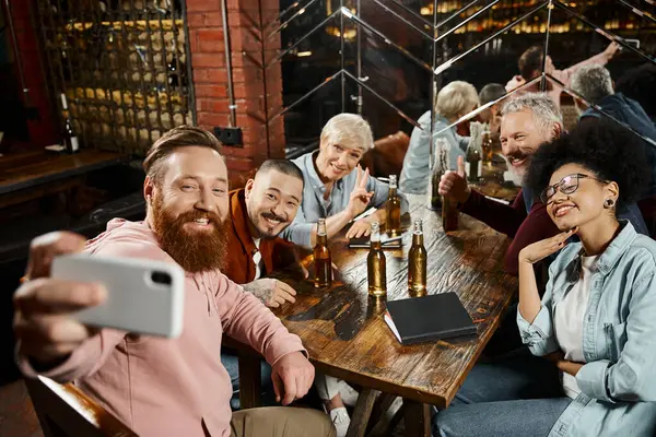 Happy bearded man taking photo with multiethnic workmates near beer bottles on wooden table in bar — Stock Photo