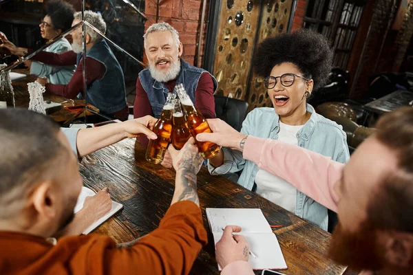 Overjoyed multiethnic workmates clinking beer bottles while resting in pub after work, party time — Stock Photo