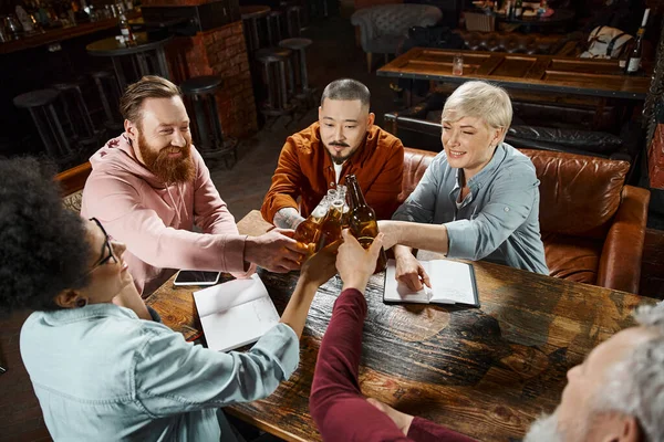 Alegres colegas multiculturales tintinean botellas de cerveza cerca de los cuadernos en la mesa de madera en el pub - foto de stock