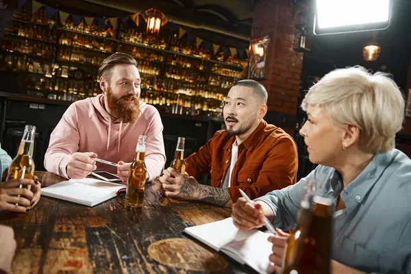 Tattooed asian man discussing startup with diverse team near beer bottles during meeting in pub — Stock Photo