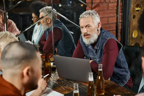 Бородатий чоловік сидить біля ноутбука і обговорює стартап-проект з багатоетнічною командою в барі — стокове фото