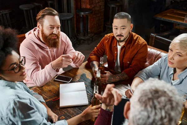 Colegas multiétnicos sentados en el pub y discutiendo el proyecto de negocios cerca de botellas de cerveza y portátil - foto de stock