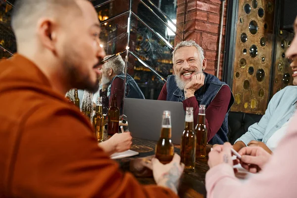 Щасливий бородатий чоловік посміхається біля ноутбука і розмиті багатоетнічні друзі розмовляють в пабі після роботи — стокове фото