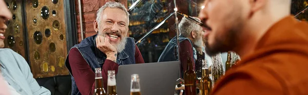 Щасливий бородатий чоловік сидить поруч з ноутбуком і багатоетнічними роботодавцями, розмовляючи в пабі після роботи, банер — стокове фото