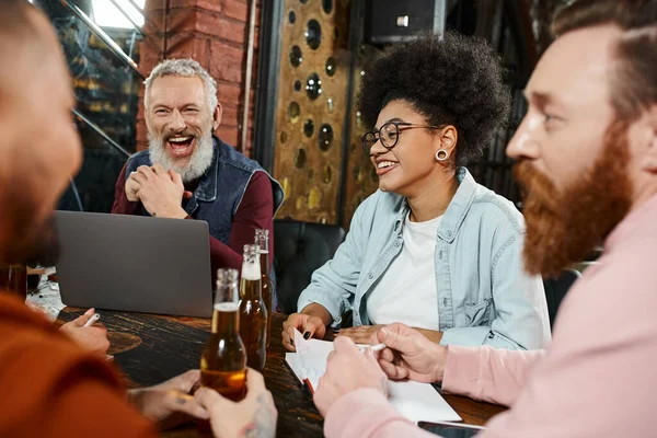 Збуджений бородатий чоловік сміється біля ноутбука та багатоетнічних робочих сусідів з пляшками пива, що розмовляють у пабі — стокове фото