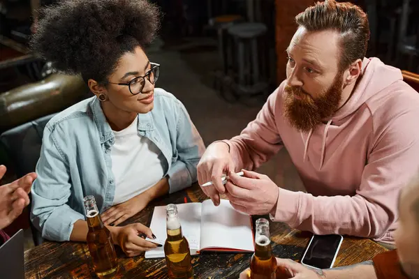 Homem barbudo conversando com equipe multiétnica enquanto sentado no pub perto de notebook e garrafas de cerveja — Fotografia de Stock