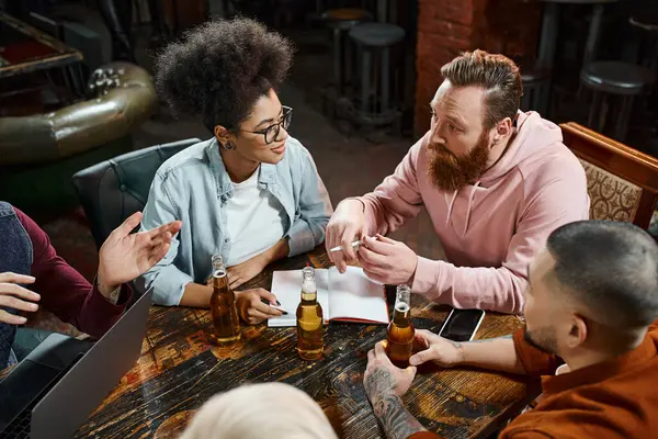 Diverse multiethnic team talking near devices and beer bottles while sitting in pub after work — Stock Photo