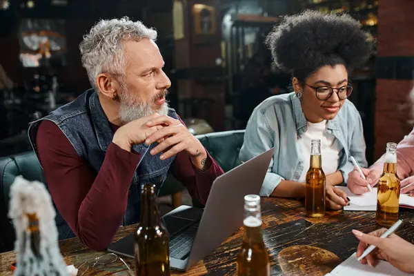 Hombre barbudo escuchando a colegas multiétnicos cerca de la computadora portátil y la cerveza en el pub, el trabajo y el ocio - foto de stock