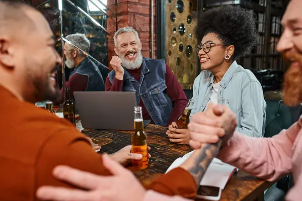 Веселий бородатий чоловік дивиться на багатоетнічних колег, тремтячи руками в барі біля пляшок пива — стокове фото