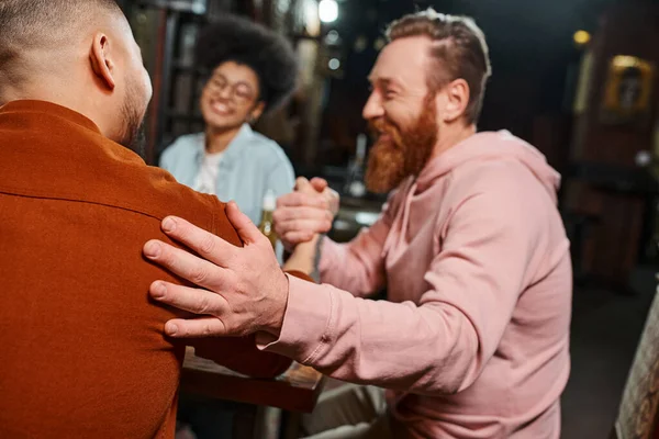 Homem barbudo satisfeito apertando as mãos com colega no pub perto de mulher americana africana borrada — Fotografia de Stock