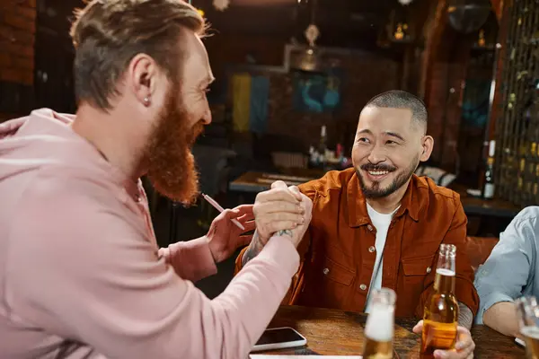 Радісний азіатський чоловік тремтить руками з бородатим колегою, закриваючи угоду поблизу пляшок пива в пабі — стокове фото
