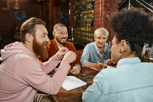 Equipo multiétnico feliz sentado en el pub cerca de cerveza y cuadernos mientras discute el proyecto de inicio - foto de stock