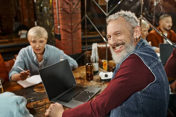 Homem barbudo alegre olhando para a câmera perto de laptop e colegas multiétnicos planejamento projeto no pub — Fotografia de Stock