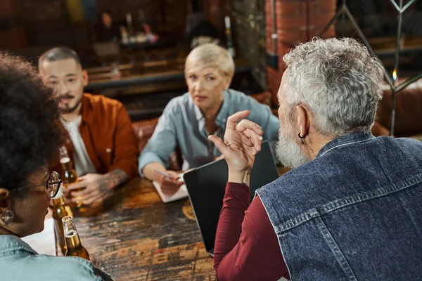 Hombre de mediana edad hablando con colegas multiétnicos atentos mientras planea un nuevo proyecto en el pub - foto de stock