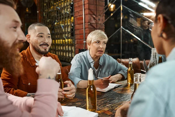 Creative multicultural team sitting in pub near beer bottles and discussing new business project — Stock Photo