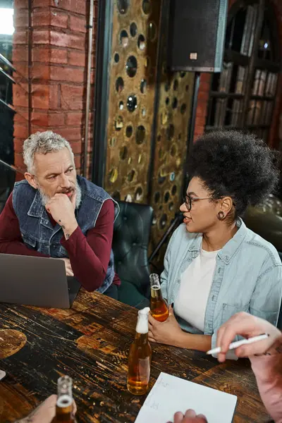 African american woman with beer bottle talking to bearded colleague sitting next to laptop in pub — Stock Photo