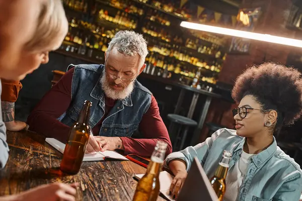 Бородатий чоловік середнього віку пише в блокноті біля пляшок пива та багатоетнічних колег у пабі — стокове фото