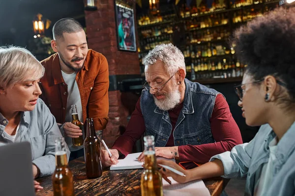 Bearded man writing in notebook and planning new startup project with multiethnic colleagues in bar — Stock Photo
