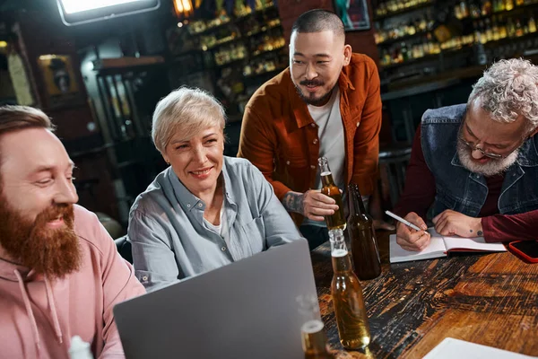 Homme d'affaires barbu écrivant dans un carnet près de l'équipe multiethnique parlant près de la bière et ordinateur portable dans un pub — Photo de stock