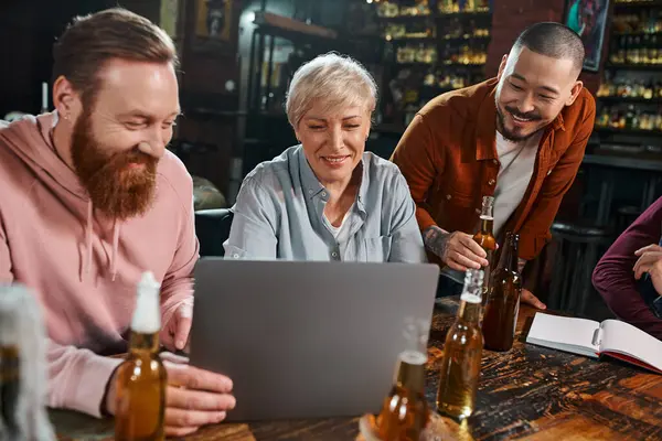 Colegas multiculturales felices mirando el ordenador portátil mientras se sienta con botellas de cerveza en el pub después del trabajo - foto de stock