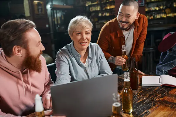 Feliz mujer de mediana edad sonriendo cerca de colegas barbudos multiétnicos proyecto de planificación en el pub - foto de stock