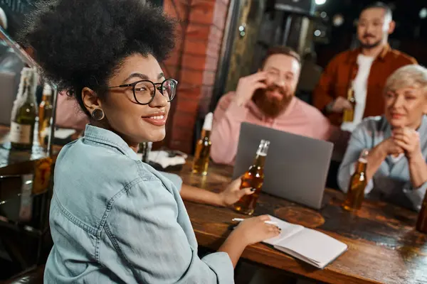 Mujer afroamericana feliz mirando a la cámara cerca de equipo multiétnico sobre fondo borroso en el bar - foto de stock