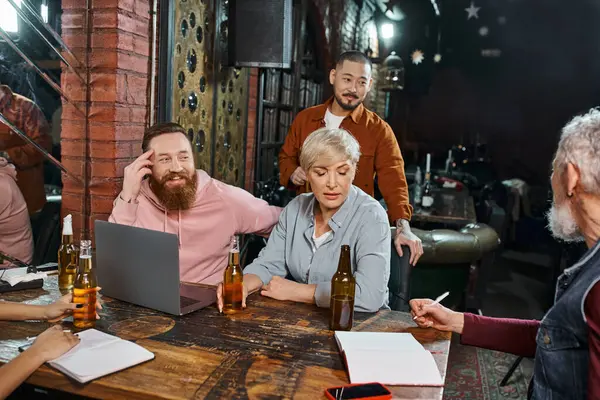 Equipo multicultural sentado en el pub cerca de botellas de cerveza y portátil y discutir nuevo proyecto de negocio - foto de stock