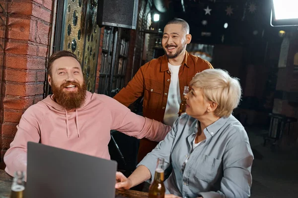 Hombre barbudo alegre sonriendo cerca de la computadora portátil y colegas multiétnicos hablando en el pub después del trabajo - foto de stock