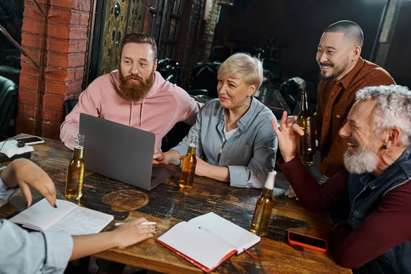 Cheerful multiethnic workmates listening to colleague near laptop and beer bottles in pub after work — Stock Photo