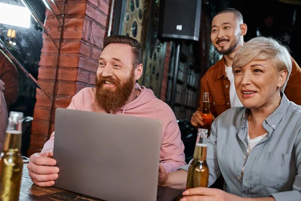 Cheerful multiethnic team smiling near laptop and beer bottles while spending time in pub after work — Stock Photo