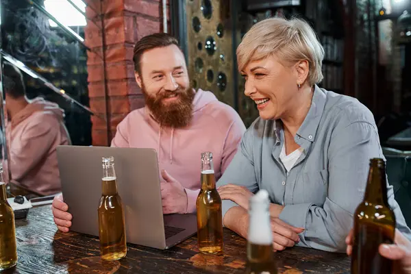 Uomo barbuto sorridente parlando con allegro collega di mezza età vicino a bottiglie di birra e laptop in pub — Foto stock